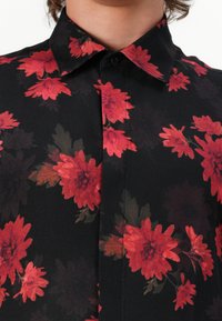 Sort skjorte med knapper og blomsterdesign, der har røde og mørkebordeaux blomster. Stoffet har en glat tekstur og en blød finish.