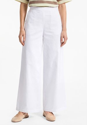 Personne portant un pantalon blanc taille haute à jambes larges associé à des chaussures plates beige et un haut à manches courtes rayé beige et marron.