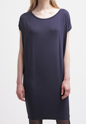 Kobieta ubrana w luźną, krótkorękawową, ciemnogranatową sukienkę do kolan na jasnym, jednolitym tle.