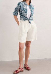 Chemise à motifs floraux bleus, shorts en lin blancs, et sandales rouges à lanières. La chemise présente des manches roulées et un devant noué.