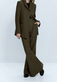 Costume ajusté vert olive avec une veste à un bouton, dotée d'une poche avant et d'un pantalon large. Tissu lisse avec une texture subtile.