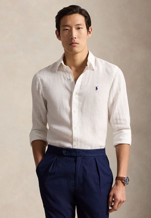 Man die een wit linnen overhemd met knopen en opgerolde mouwen draagt, marineblauwe geplooide broek en een bruin polshorloge, staand tegen een effen achtergrond.