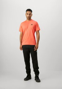Camiseta deportiva naranja con cuello redondo y logotipo negro de Adidas, combinada con pantalones de chándal negros y zapatillas negras, sobre un fondo liso.