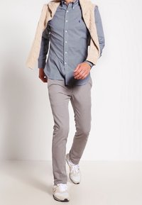 Man som går och bär en ljusblå skjorta, beiga byxor, vita sneakers och en beige tröja över axlarna.