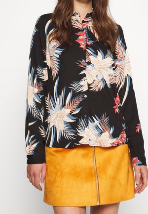 Chemise à manches longues en tissu noir avec un motif floral vibrant en crème, bleu et rouge, associée à une jupe en daim jaune à fermeture éclair sur le devant.