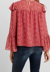 Rød blomsterbluse med flæser på ærmerne og nøglehuldetaille i ryggen. Letvægtsstof med et subtilt mønster, parret med mørke jeans.