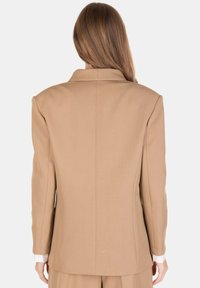 Blazer beige su misura con spalle strutturate, dettagli sulla cucitura posteriore e colletto. Indossato sopra una camicia bianca con i polsini visibili.