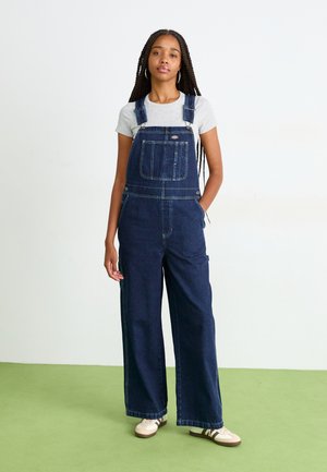 Salopette in denim blu scuro, con gambe larghe, tasca frontale e spalline regolabili, abbinata a una maglietta grigia chiara a maniche corte e sneakers.