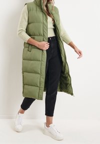 Dlouhá polstrovaná olivově zelená vesta s vysokým límcem, předními knoflíky a bočními kapsami, nošená přes světle zelený pletený svetr a černé džíny.