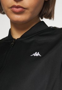 Gros plan sur une personne portant une veste noire zippée avec un logo blanc représentant deux figures assises sur la poitrine.