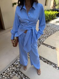 Conjunto de dois peças azul listrado, composto por uma camisa de botão com um laço na cintura e calças largas, ambos adornados com pequenos detalhes em ouro.