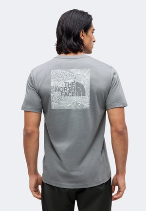 Mężczyzna o ciemnych włosach, ubrany w szarą koszulkę z dużym białym nadrukiem i logo The North Face na plecach, stojący na jednolitym tle.