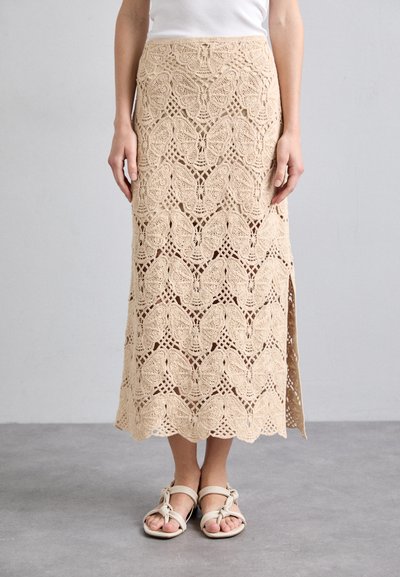 Jupe en crochet beige avec un motif de papillon, dotée d'une fente sur le côté et d'un design texturé. Associée à des sandales de couleur claire.