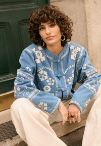 Ženska z kodrastimi lasmi, ki nosi okrogle uhane, modro jakno z rožnatim vzorcem in kremne hlače, sedi zunaj ob zelenih vratih.