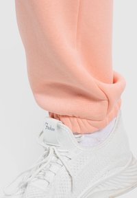 Peachfarvede sweatpants med manchetter ved anklerne, parret med hvide mesh sneakers. Bukserne har en glat, tætsiddende tekstur.