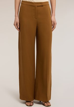 Femme portant un pantalon marron taille haute à jambes larges, associé à des sandales noires ouvertes aux orteils, debout devant un fond beige neutre.