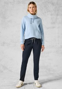 Ljust blå hoodie med en frampocket, tillsammans med marinblå joggerbyxor med dragsko i midjan och sidfickor. Vita sneakers.