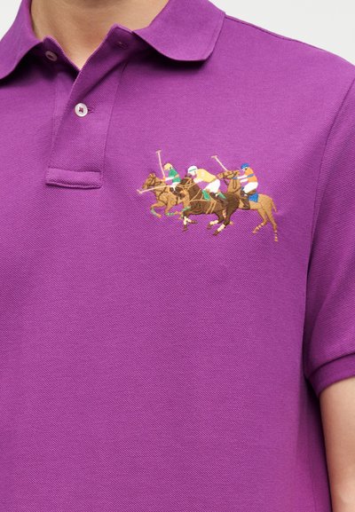 Μωβ πόλο μπλούζα με κεντημένο λογότυπο τριών παίκτών πόλο πάνω σε άλογα σε δράση κοντά στο κολάρο στη αριστερή πλευρά.