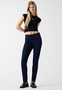 Μαύρο κοντομάνικο κοντό τοπ, σε συνδυασμό με σκουρό μπλε στενά τζιν. Το τζιν έχει διακριτική ραφή και κλασικό κούμπωμα με κουμπί.