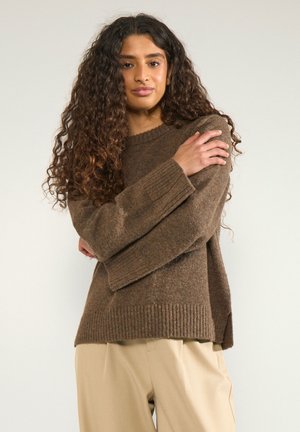 Femme aux longs cheveux bouclés portant un pull en maille marron et un pantalon beige, debout, les bras croisés sur la poitrine, devant un fond clair.
