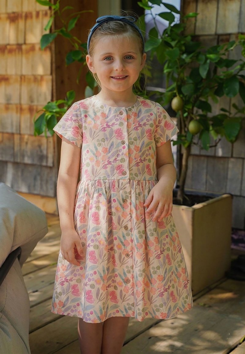 Lächendes junges Mädchen, das ein geblümtes, knöpfbares Kleid und ein blaues Stirnband trägt, steht auf einer hölzernen Veranda mit Pflanzen im Hintergrund.
