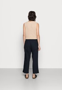 Person med kort mörkt hår iklädd en ärmlös beige stickad topp, svarta lösa byxor och svarta sandaler, stående vänd mot en helt vit vägg.