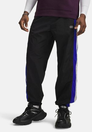 Man wearing black athletic pants with blue and white side stripes and black sneakers with white accents against white background.