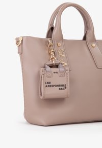Beige leren handtas met gouden hardware, voorzien van een kleine afneembare zak met het label "IK BEN EEN VERANTWOORDELIJKE TAS." Gladde textuur, gestructureerd ontwerp.