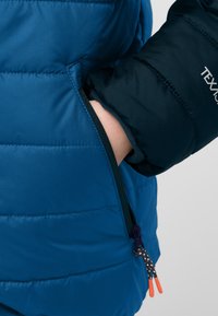Giacca puffer blu con una tasca laterale con cerniera. Il materiale ha una superficie liscia, con maniche blu scuro a contrasto e tirazip arancione.