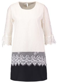 Robe en dentelle blanche avec manches trois-quarts, rubans noués aux poignets, fermeture à bouton avec trou en forme de goutte dans le dos, et un ourlet noir avec des détails en dentelle.