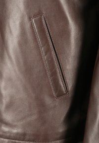 Brun läderjacka med en slät textur, med en horisontell dragkedjeficka på sidan, som betonar rena linjer och minimalistisk design.