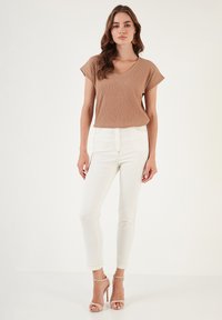 Light brown ribbed short-sleeve top paired with white fitted jeans, standing model wears beige strappy heels against a plain background.