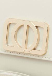 Bílá kožená kabelka s pozlaceným dvojitým kruhem a obdélníkovým zapínáním, hladkém povrchu a čistém designu.