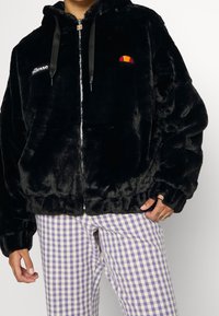 Sort faux fur jakke med lynlås fortil, justerbare sorte satin snore og broderet logo. Sammen med lilla ternede bukser.