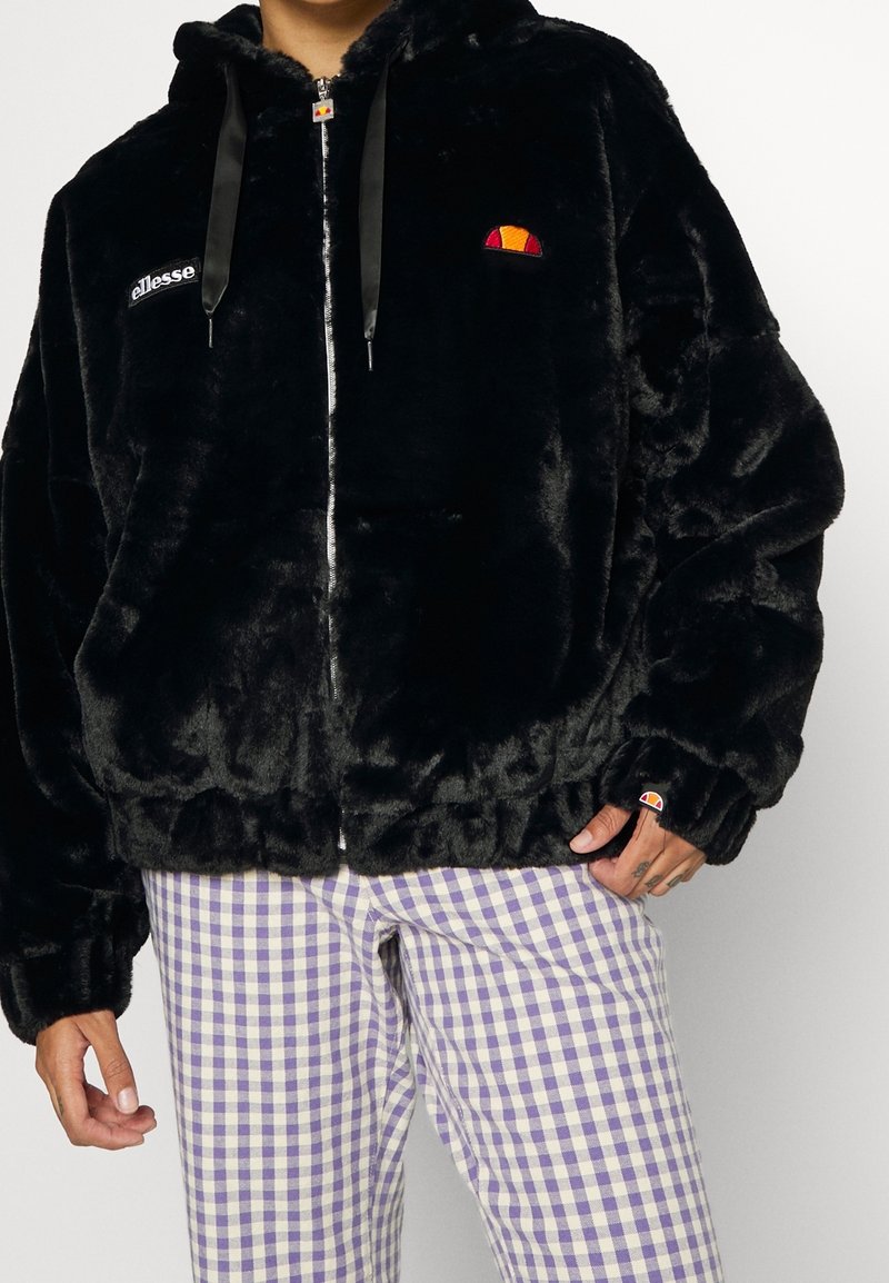 Sort faux fur jakke med lynlås fortil, justerbare sorte satin snore og broderet logo. Sammen med lilla ternede bukser.
