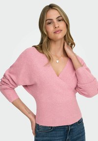 Różowy sweter z dzianiny z kopertowym dekoltem w kształcie litery V, długimi rękawami i prążkowanym dołem. Miękka faktura i luźny krój, zestawiony z jeansami.