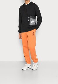 Černá dlouhá mikina, oranžové tepláky s logem, šedá křížová taška a bílé tenisky s černými akcenty. Jednoduchý, sportovní outfit.