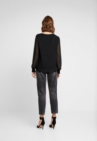 Svart blus med genomskinliga långa ärmar, rund halsringning och elastiska manschetter. Kombinerad med grå denimbyxor och svarta högklackade sandaler.