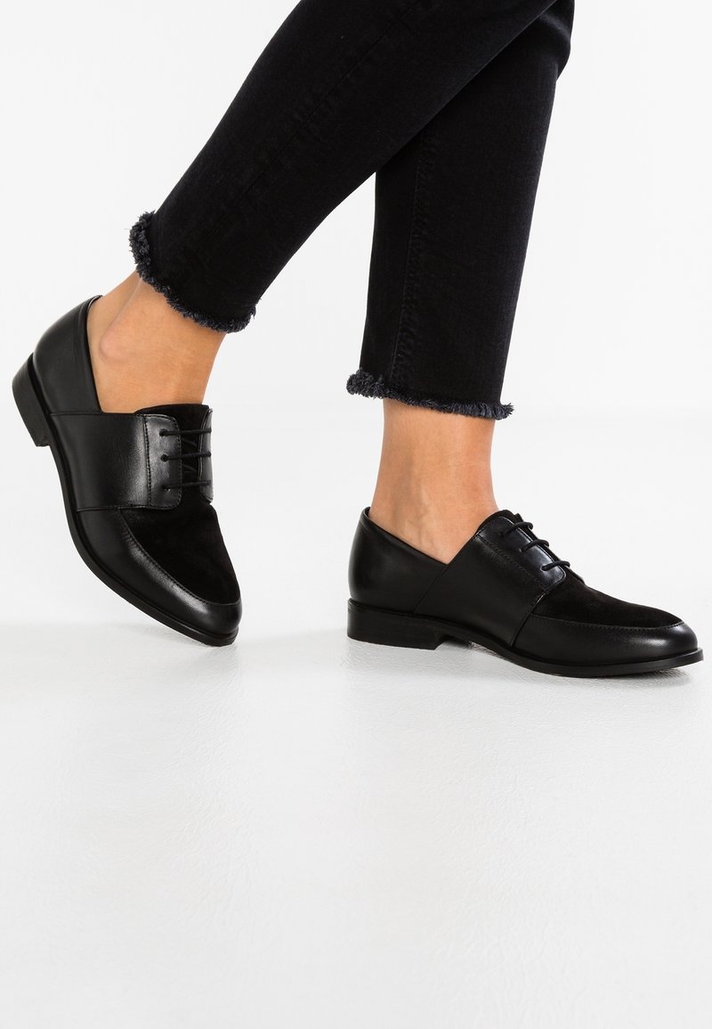 Chaussures en cuir noir et en daim avec un détail à lacets et un bout arrondi, portées avec un jean noir court avec des ourlets effilochés.