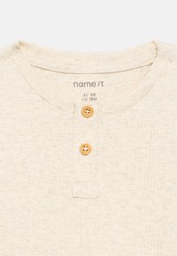 Φωτεινό μπεζ ριγωτό βαμβακερό πουκάμισο με στρογγυλή λαιμόκοψη και δύο ξύλινα κουμπιά στην πατιλέτα. Η ετικέτα γράφει "name it", μέγεθος EU 86, US 18M.
