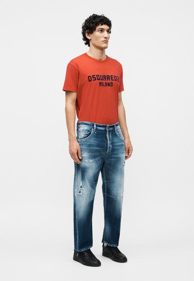 Άνδρας με σγουρά μαλλιά που φοράει κόκκινη μπλούζα DSQUARED2 Milano, ξεφτισμένο μπλε τζιν και μαύρα αθλητικά, στέκεται μπροστά σε λευκό φόντο.