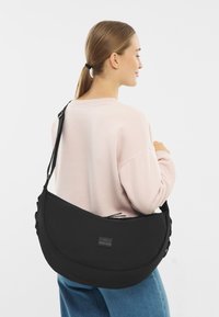 Czarna torba na ramię z zaokrąglonym kształtem, z zapięciem na zamek i naszywką z logo, noszona na ramieniu. Gładka faktura.
