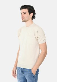 Maglione a maniche corte beige in maglia, con scollatura rotonda e taschino sul lato sinistro del petto. Texture liscia con orlo e bordi delle maniche a costine. Vestibilità casual.