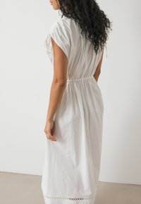 Robe en coton blanc avec des manches courtes, taille froncée et dentelle au niveau de l'ourlet. Texture lisse et coupe décontractée. Vue de côté affichée.