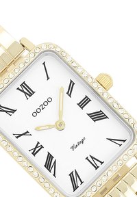 Montre rectangulaire dorée avec des chiffres romains, cadran blanc et accents en cristal autour de la lunette. Bracelet en métal avec finition polie.