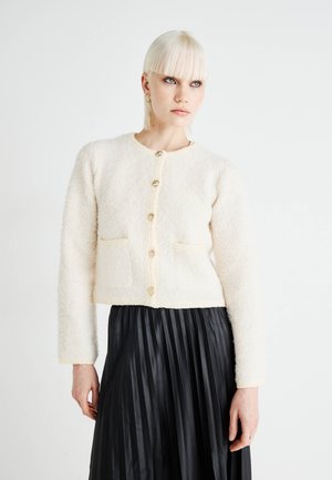 Ivory textured cardigan with a round neckline, front pockets, and button closure. Styled over a pleated black skirt, featuring long sleeves.