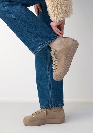 Personne portant un jean bleu et des baskets beiges ajuste le ourlet en se tenant debout sur un sol gris clair sur un fond uni.