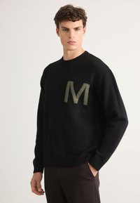 Suéter negro de punto con cuello redondo, que presenta una "M" verde texturizada en el pecho. Puños y dobladillo acanalados. Acabado de tela suave.