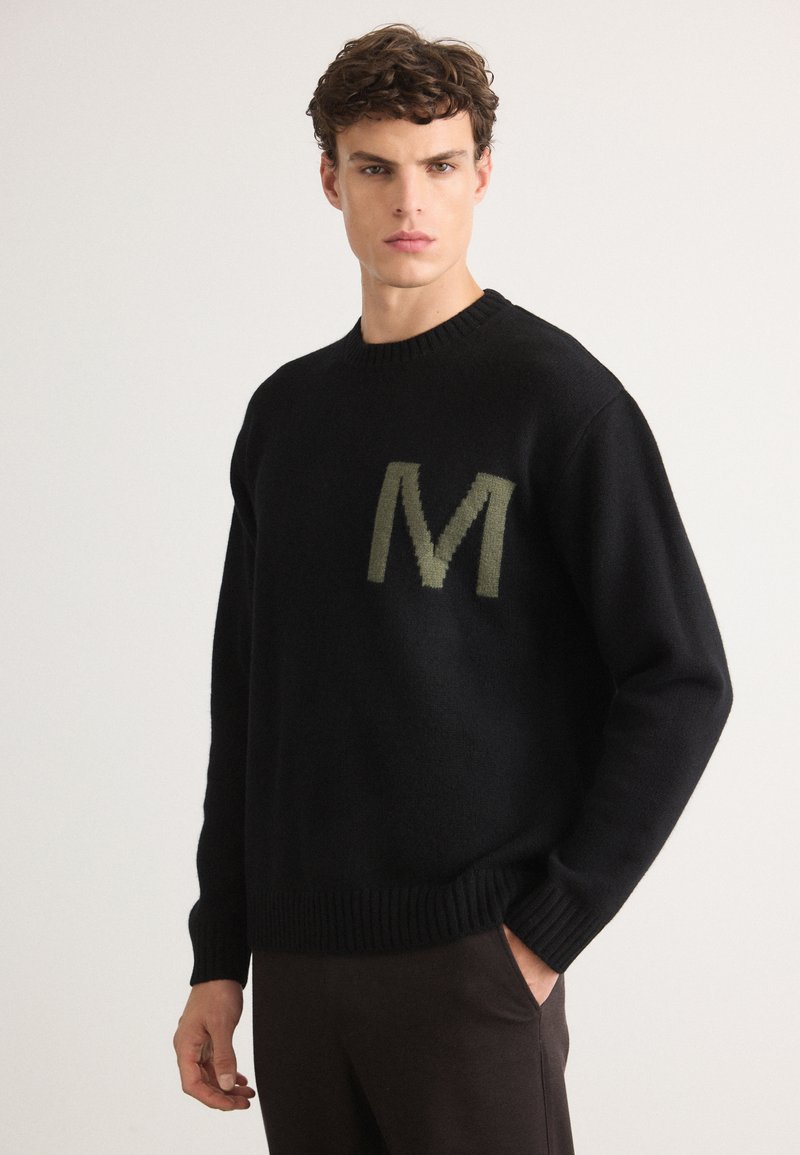 Suéter negro de punto con cuello redondo, que presenta una "M" verde texturizada en el pecho. Puños y dobladillo acanalados. Acabado de tela suave.