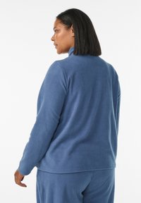 Sininen fleece-paita korkealla kauluksella, sileä tekstuuri ja pitkät hihat. Näkyvät saumat ja rento istuvuus.